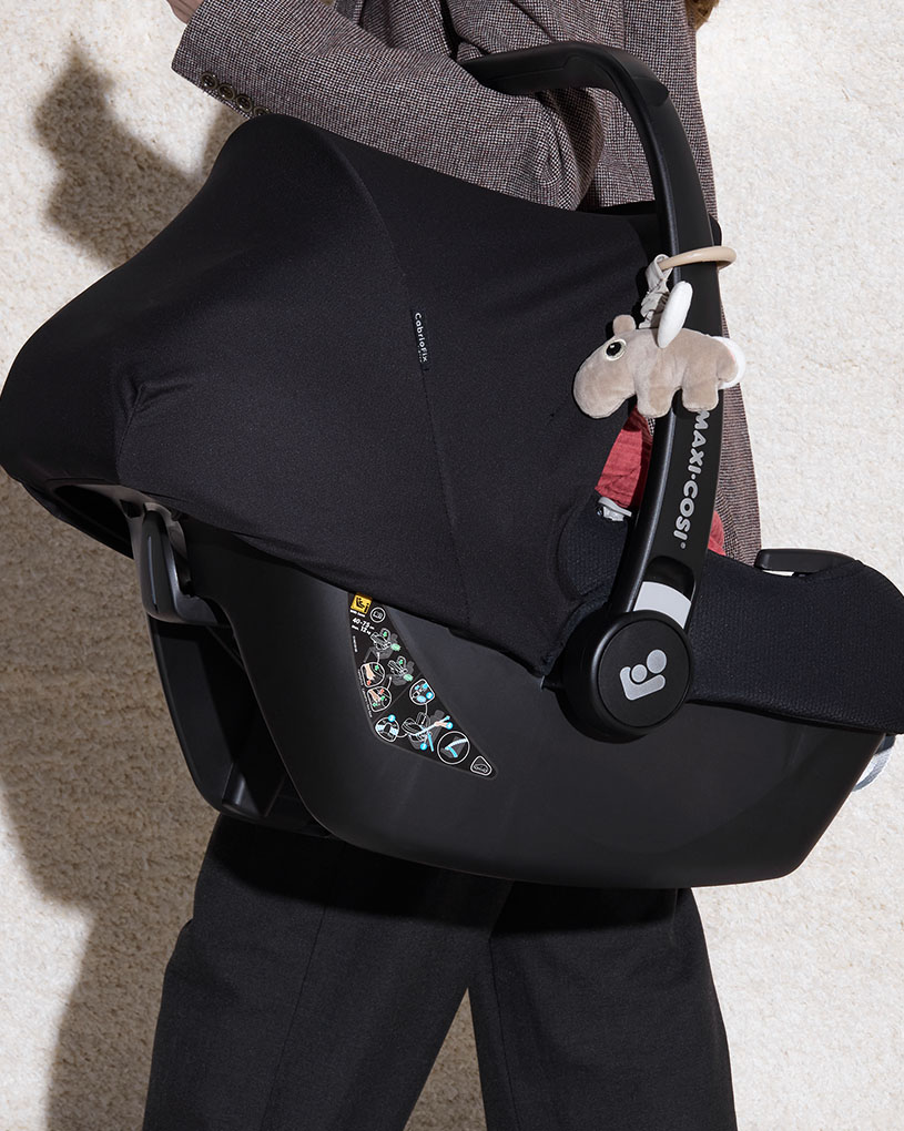 En person bär ett babyskydd med en leksak, med texten 'Maxi-Cosi'.